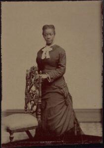 Sepia colored image of a dark-skinned woman with dark hair dress in a 19th century grown with a white collar and bow. She is standing in front of an arm chair. 