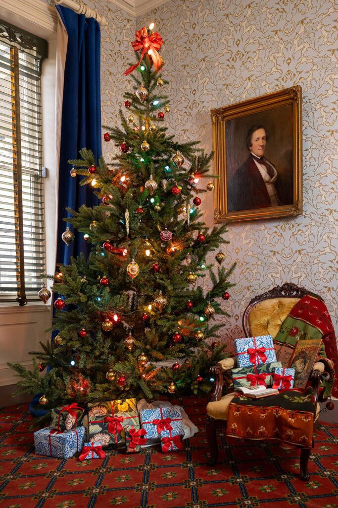A Christmas tree decorated with ornaments and lights surrounded by red-ribboned gifts in a historic parlor.