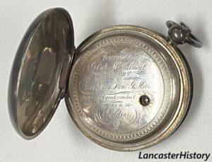 Open back cover of silver pocket watch showing second cover with inscription covering movement