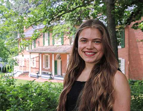 A young woman with light skin and long, wavy blond hair standing in front of a historic home.