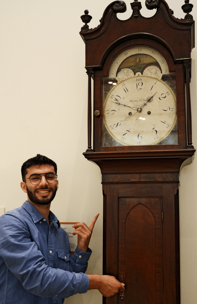 A young man gestures with a smile to a large antique clock.