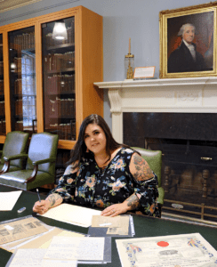 A young student sits in a room lined with books. Historic documents are scattered in front of her.