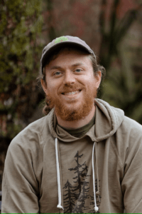 A young man with light skin and a reddish beard wearing a baseball cap and a olive colored sweatshirt.