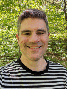 A headshot photo of a man with light skin wearing a black and white t-shirt.