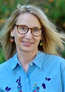 A woman with light skin and mid-length blonde hair. She wears glasses, butterfly earrings, and a blue button up shirt.