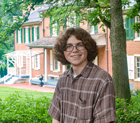 A young man with light skin and curly brown hair stands in front of a historic house.