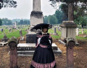 An African-American woman is dressed in period costume in front of a large gravestone.