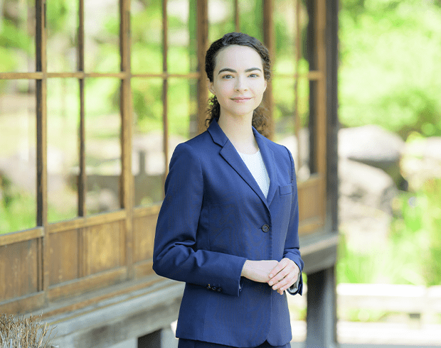 A photograph of a young woman with light skin and dark, wavy hair in a blue blazer standing in front of a Japanese style building.