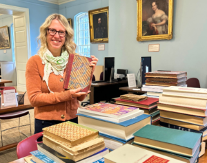 light-skinned woman with blond hair wearing a green and white scarf and rust colored sweater holds up an antique book in front of a table of books in a library.