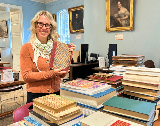 light-skinned woman with blond hair wearing a green and white scarf and rust colored sweater holds up an antique book in front of a table of books in a library.