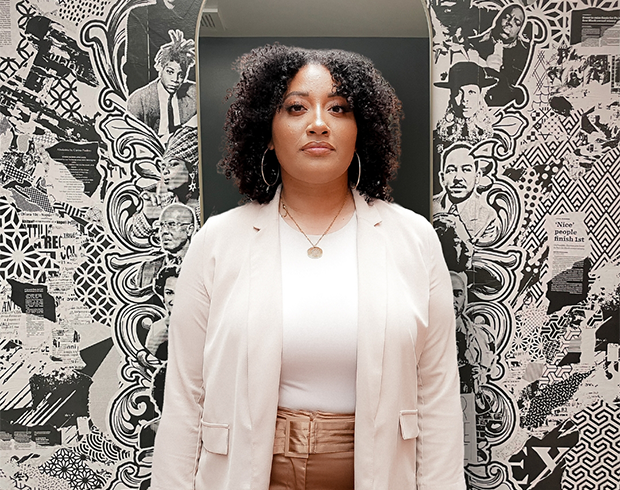A young Black woman with brown skin with brownish-black curly hair wears a cream color blazer in front of a mirror and collage background.