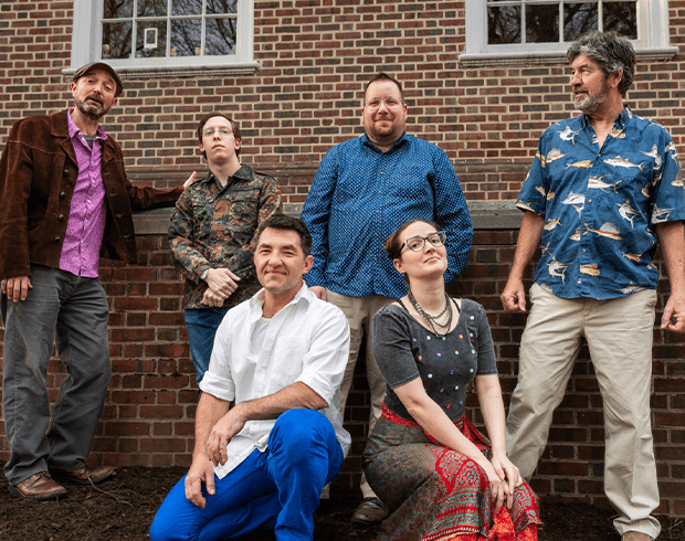 A group of six people standing, posing for the camera in front of a brick building.