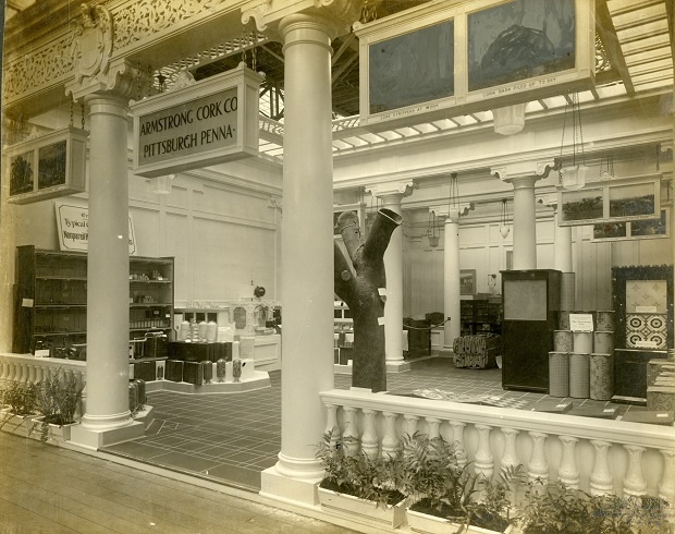 A historic photograph of the interior of the Armstrong Pavilion. Ionic columns flank the entrance with a sign saying "Armstrong Cork Company, Pittsburgh, PA." The interior features different displays and wood furnishings.