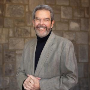 A headshot of a man with light skin. He wears a black turtleneck shirt with a gray blazer on top. His hands are clasped in front of him and he smiles at the camera.