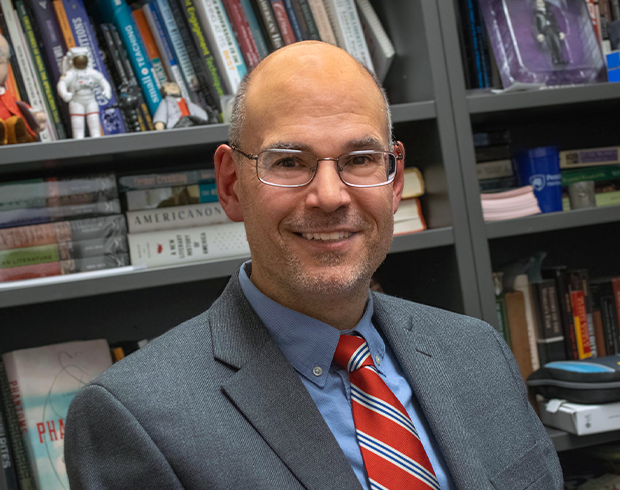 Image of an older man with light skin. He had a bad head, wears glasses, and is wearing a gray suit with a red striped tie.