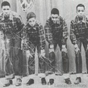 A black and white historic photograph of four young African American boys with their hands on their knees. They wear identical outfits.