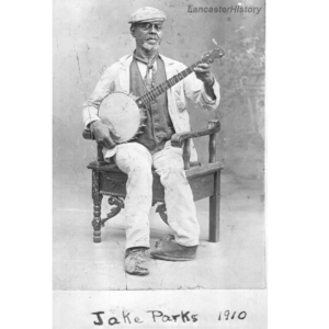 A black and white historic photo of a Black man sitting on a chair with a banjo in his hands. 