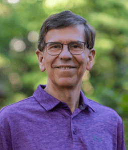 An older man with light and brown hair. He wears glasses and a purple polo shirt.