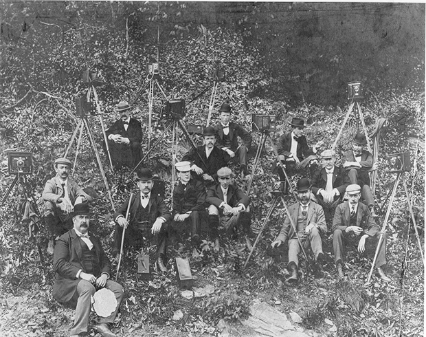 Historic image of a group of 13 men from the Lancaster Camera Club sitting on a small wooded hillside with their cameras and tripods.