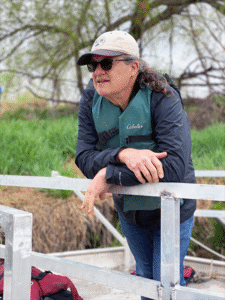 A man with light skin leans on a railing outdoors. He wears a life-vest, a hat, and sunglasses.