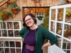 A young woman with light skin and short brown hair. She wears glasses and a purple dress with a green cardigan in front of a greenhouse.