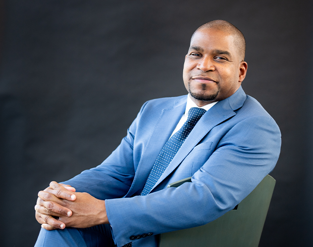 Image of a young Black man with dark skin sitting on a chair, hands crossed over his knees. He wears a light blue suit and tie.