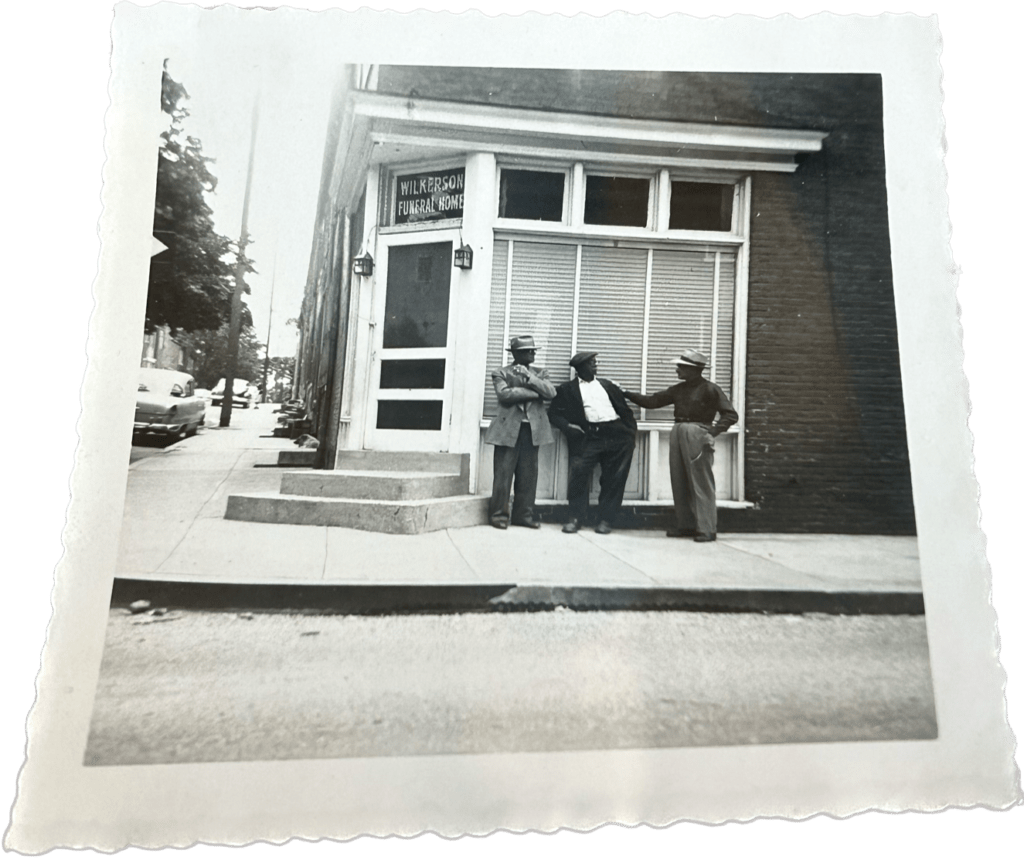 A black and white historic photo of a building with its entrance on the corner. Three Black men in suits stand outside the windows in to the funeral home in conversation.