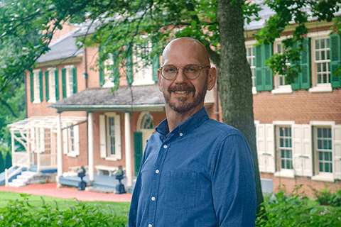 A young man with light skin, bald head, a beard, glasses, and a blue button up shirt stands in front of a historic house.