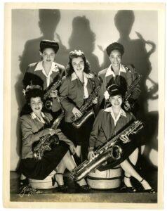 A black and white historic photograph of five women holding various saxophones.