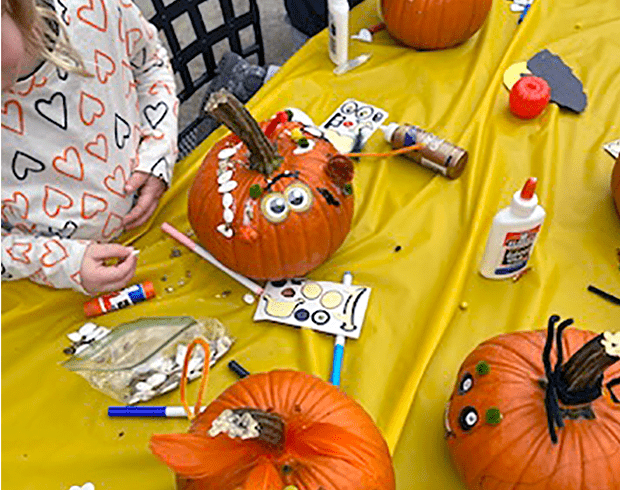A table with a bright yellow table cloth, covered in crafting supplies and pumpkins in various stages of being decorated with eyes, stickers, and puff balls.
