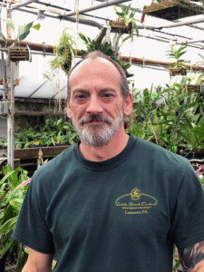 A photo of an older man with light skin and a gray beard. He is wearing a green t-shirt and standing in a greenhouse.