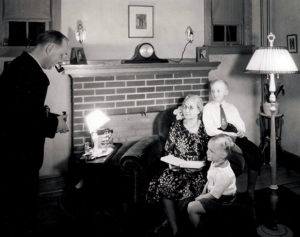 A black and white image from the mid-1900s with a woman and her two children sitting on an armchair, photographed by a man holding a camera. 