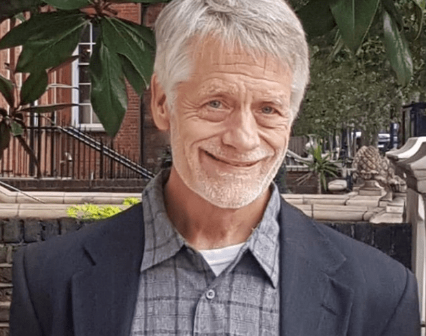 Headshot photograph of an older man with light skin. He has gray-white hair and a closely-kept beard.