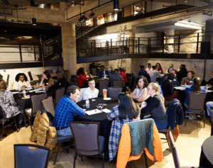 A group of people gather around multiple tables in discussion.