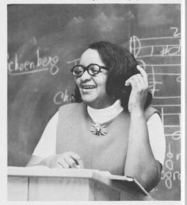 A black and white historic photograph of Romayne Bridgett standing at a lectern in front of a blackboard.