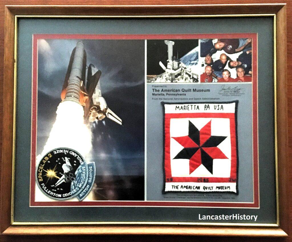 A plaque containing a miniature quilt, several photos, and a Space Shuttle Challenger mission patch.