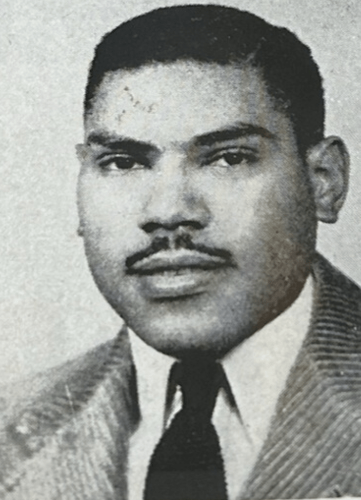 A black and white image of a Black man with black hair and a black moustache. He's looking at the camera and wearing a collared shirt, tie, and suit jacket.