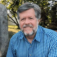 A photo of Ben Webber, a man with short gray hair and a bear wearing a blue checkered shirt.