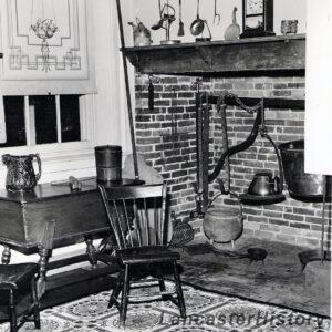 Historic photo of the Butler's Pantry interior after the Sons of the American revolution project was completed in the early 1940s. The fireplace is made of open brickwork with a large metal rail inserted in the center. On the rail is various pots used for cooking over an open flame. At the top of the fireplace is a mantle with a clock sitting upon it. Above the mantle is a pegboard with cooking utensils hanging from the pegs. To the right of the fireplace is a window with the blind pulled halfway down. At the base of the window is a small wooden worktable with two jugs sitting on top. A wooden chair sits in front of the worktable.