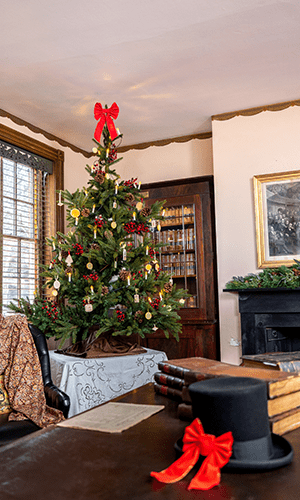 The Library in James Buchanan's Wheatland home. A top hat with a red bow sits on a table in the foreground and a table-top tree, alight with electric candles, is in the background.
