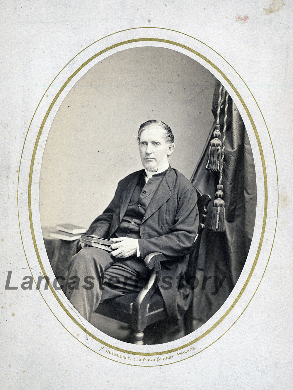 A black and white photograph of a light skinned, older adult man. He is seated in a wooden hair with his legs crossed and books in his lap. He wears a dark colored suit with a clerical collar.