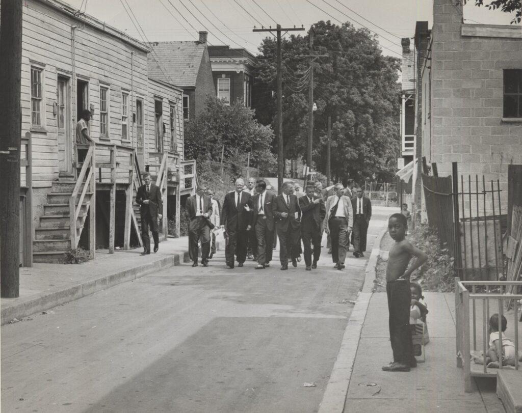 A black and white image of a residential streetscape. In the middle ground, a group of white men in suits walk down the middle of the street. A Black man and three children look on from the sides of the street. 